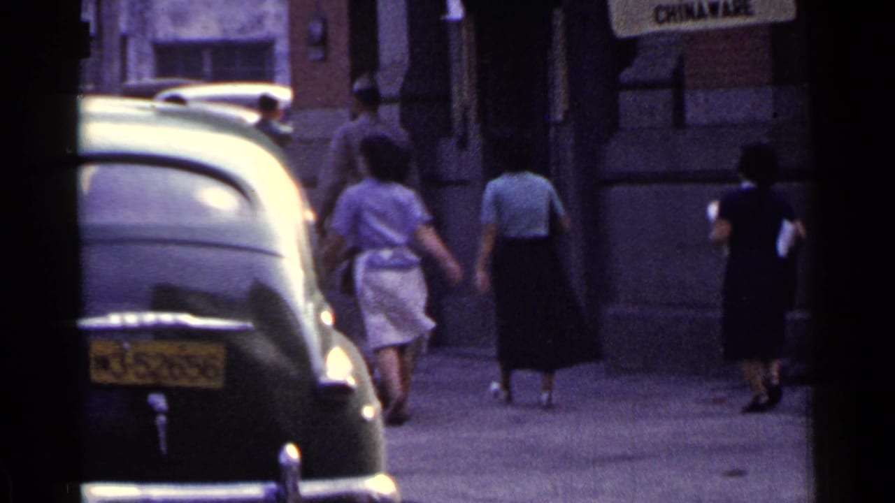 Pedestrians walk away along a sidewalk beside a dark parked sedan.