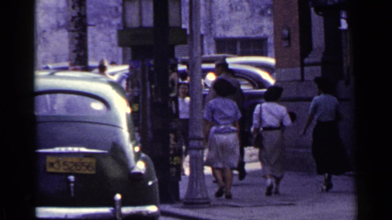 1953 Japan street view with women in dresses and other pedestrians moving away from the camera beside parked mid-century cars.