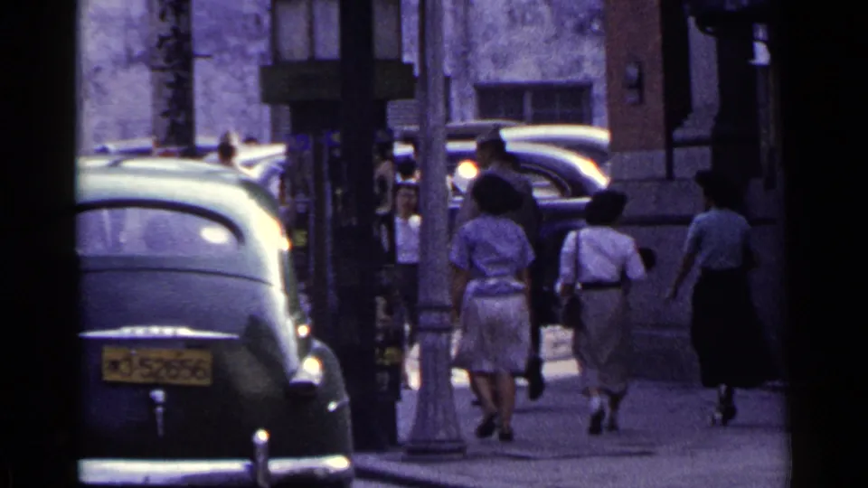 1953 Japan street view with women in dresses and other pedestrians moving away from the camera beside parked mid-century cars.