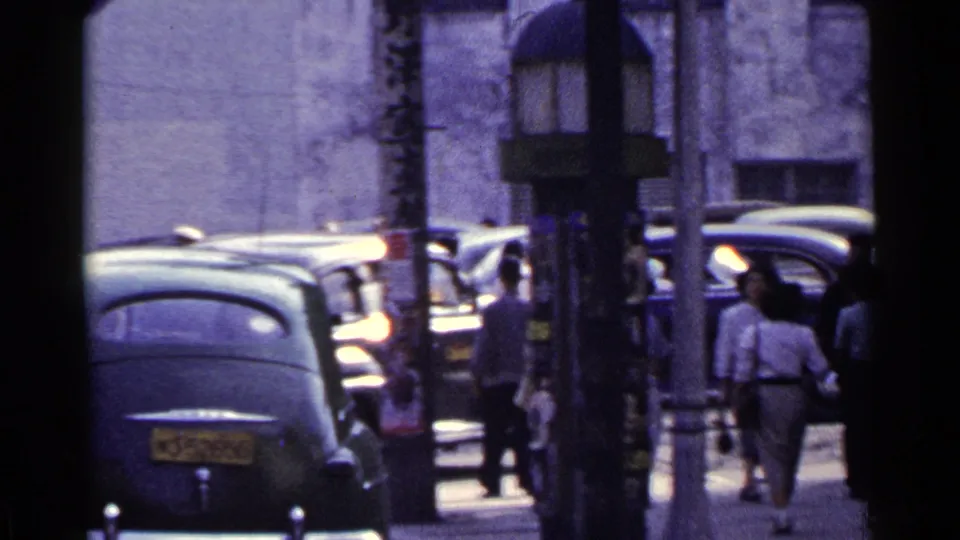 Handheld 1953 Japan frame with a dark sedan in the foreground and pedestrians moving through a narrow urban sidewalk corridor.