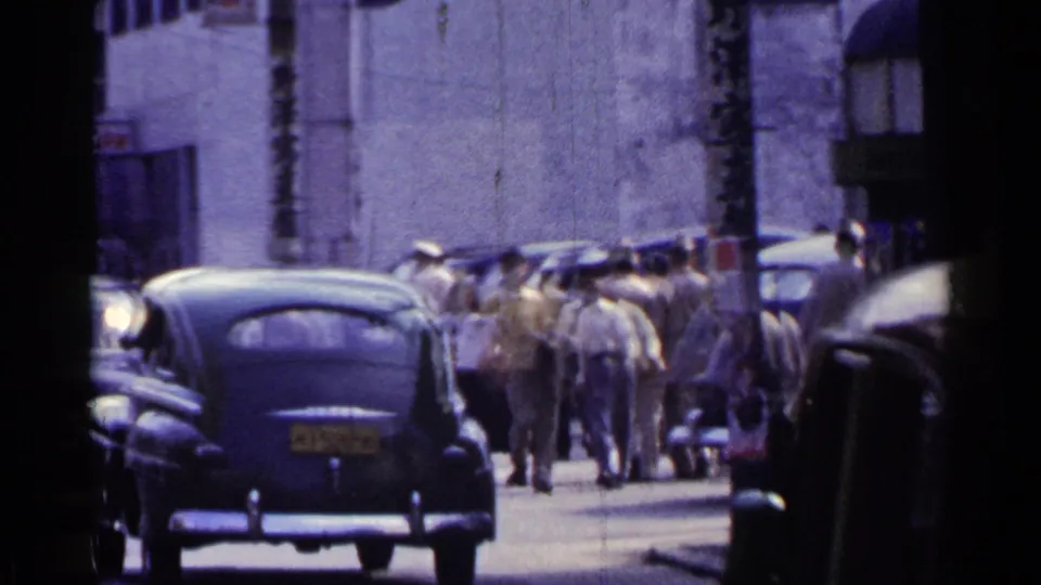 1953 Japan street frame showing parked cars, pedestrians, utility poles, and what appears to be an animal-drawn cart moving through traffic.