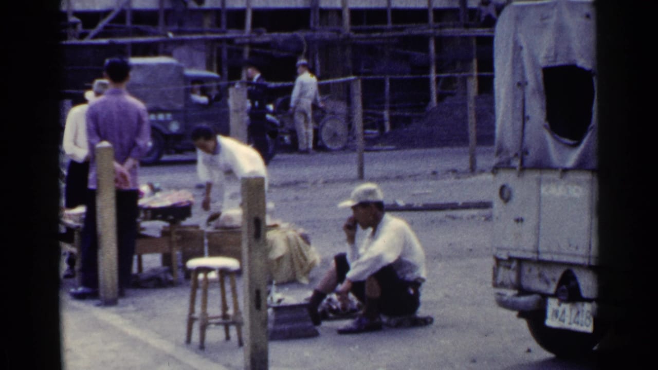 Street market view with a cobbler seated on the pavement, pedestrians, a stool, and covered vehicles behind.