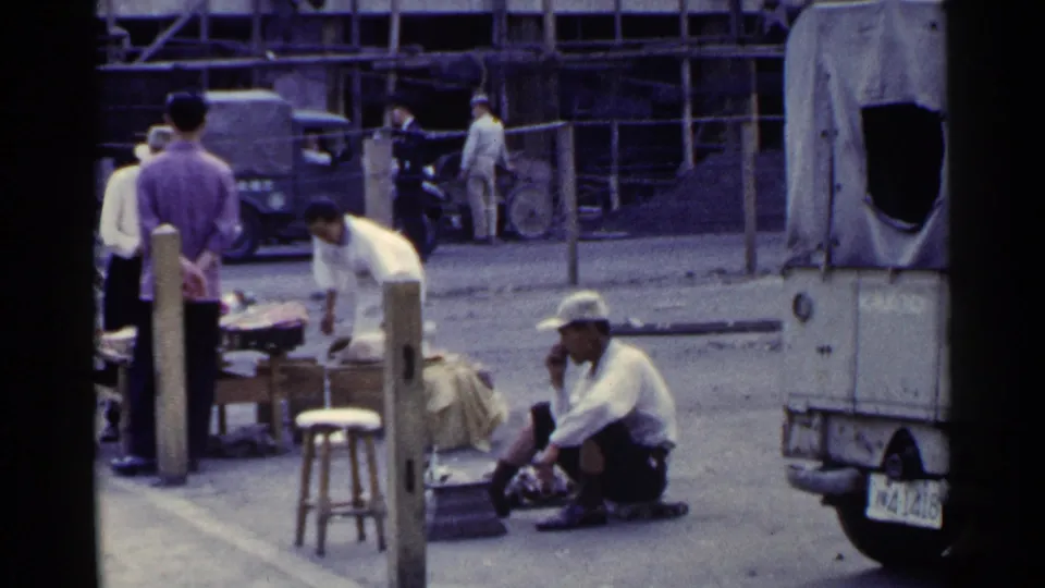 1953 Japan 8mm frame showing a cobbler working near the pavement as pedestrians stand by a small street stall with covered vehicles in the background.