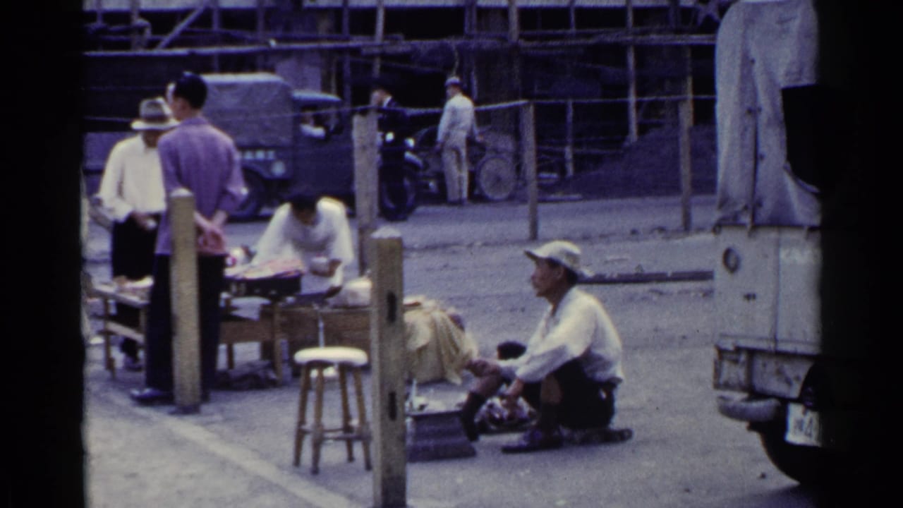 Cobbler seated by a street stall as people stand nearby and a covered truck or cart fills the right edge.