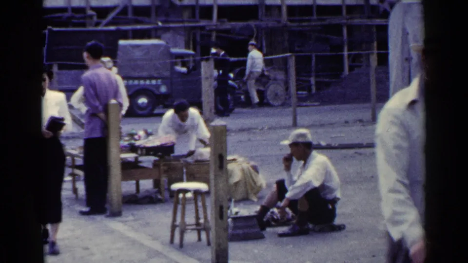 1953 Japan 8mm frame with a foreground pedestrian passing close to camera, a cobbler seated on pavement, and market activity behind roadside posts.