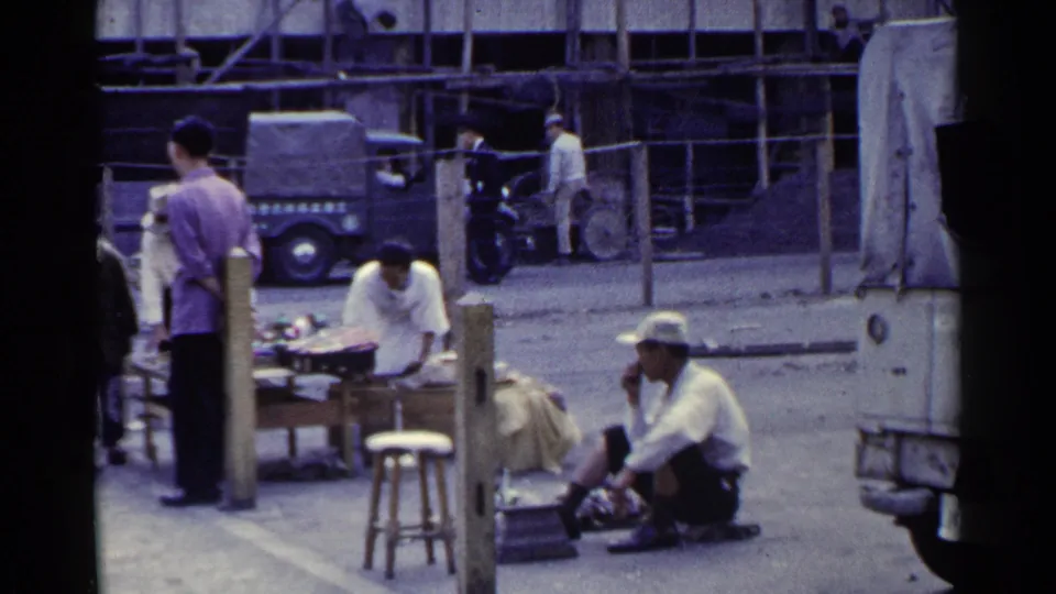 1953 Japan frame showing a cobbler at pavement level, a vendor or pedestrian at a low table, a wooden stool, covered vehicles, and roadside scaffolding.