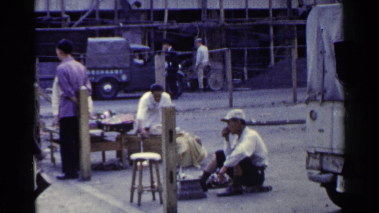 Men near sidewalk market tables with a truck and wooden buildings in the background.