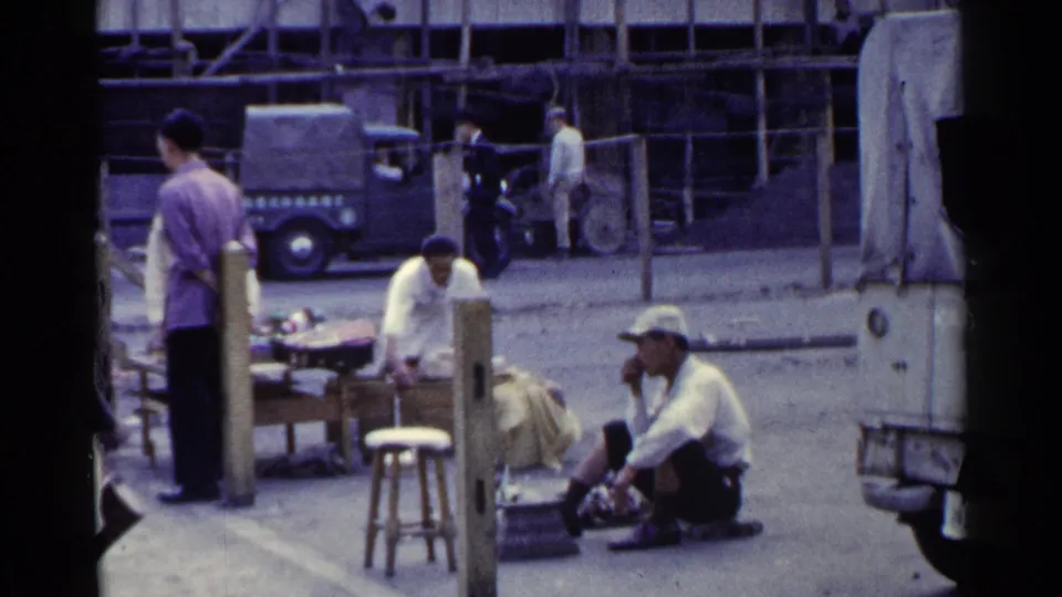 1953 Japan street-market view with men standing and crouching near tables, goods, a parked truck, and wooden storefronts.