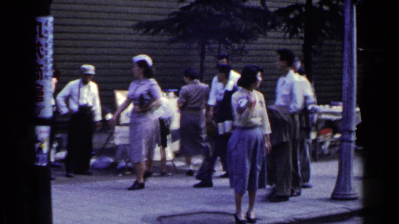 1953 Japan street scene with women and men moving near a lamppost, sidewalk stall, and shuttered building front.