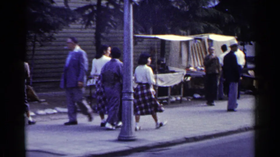 1953 Japan street-market frame with pedestrians near a canopy-covered stall, lamppost, curb, and storefronts.