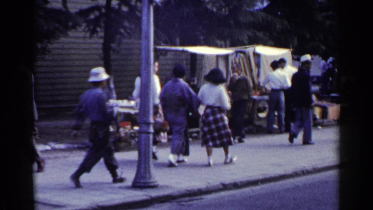 Pedestrians walk past a sidewalk stall and shoe-shine area under a canopy.