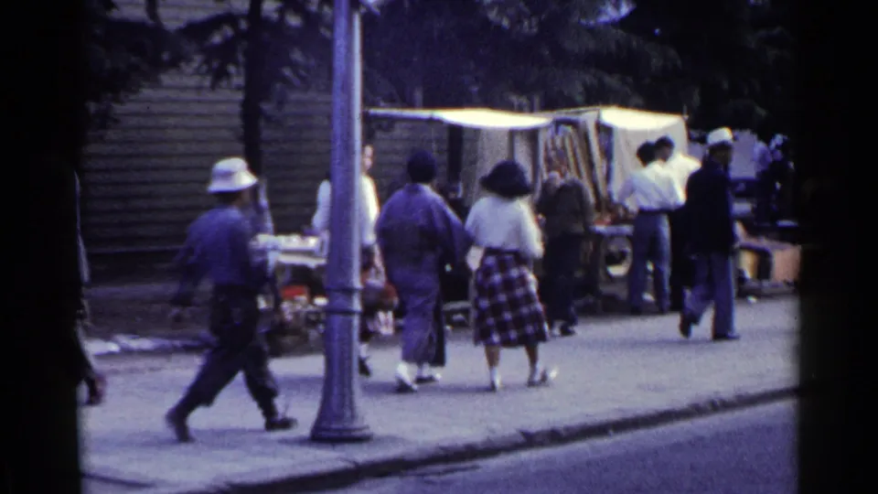 1953 Japan 8mm frame showing pedestrians passing a curbside shoe-shine station and vendor setup beneath a simple canopy.