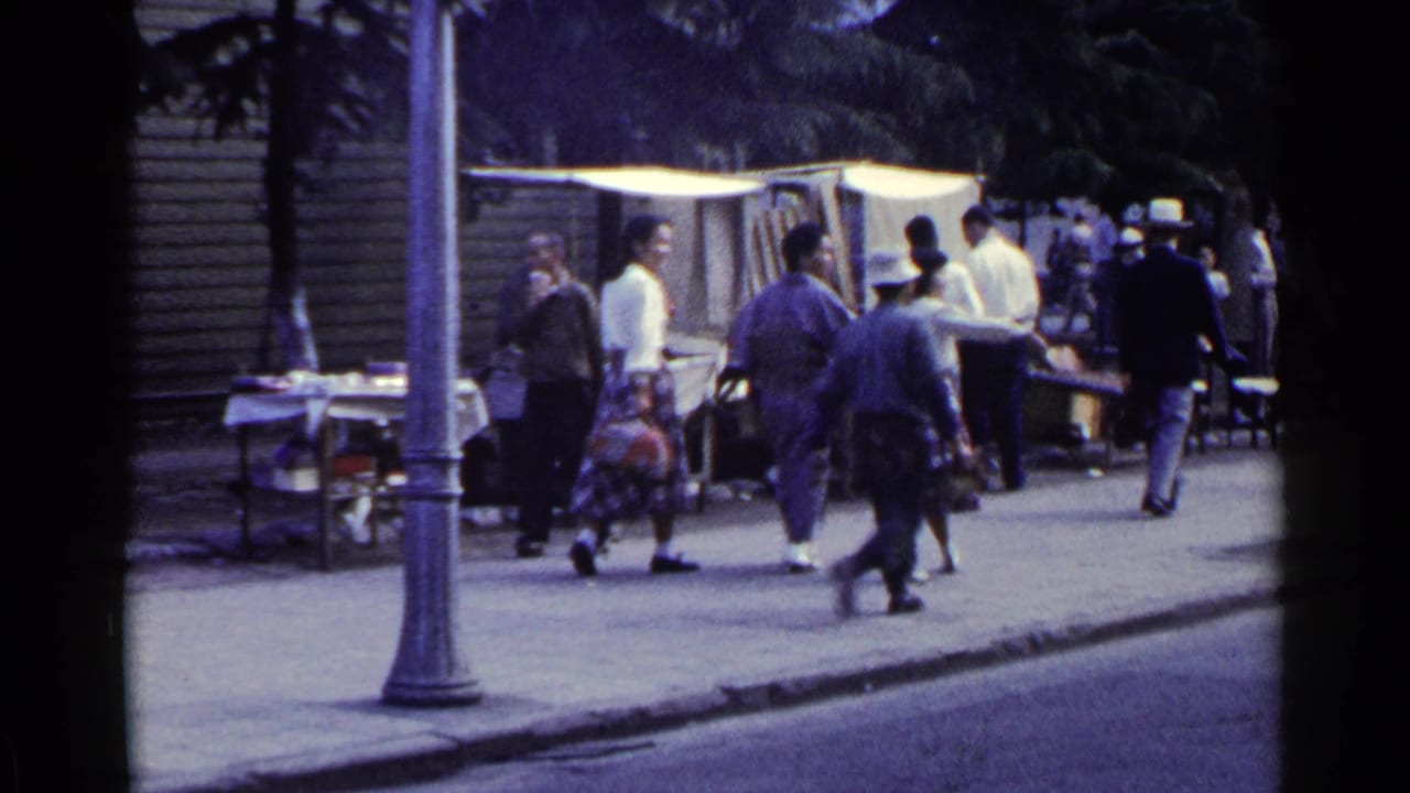People pass a lamppost beside sidewalk stalls and chairs near the curb.