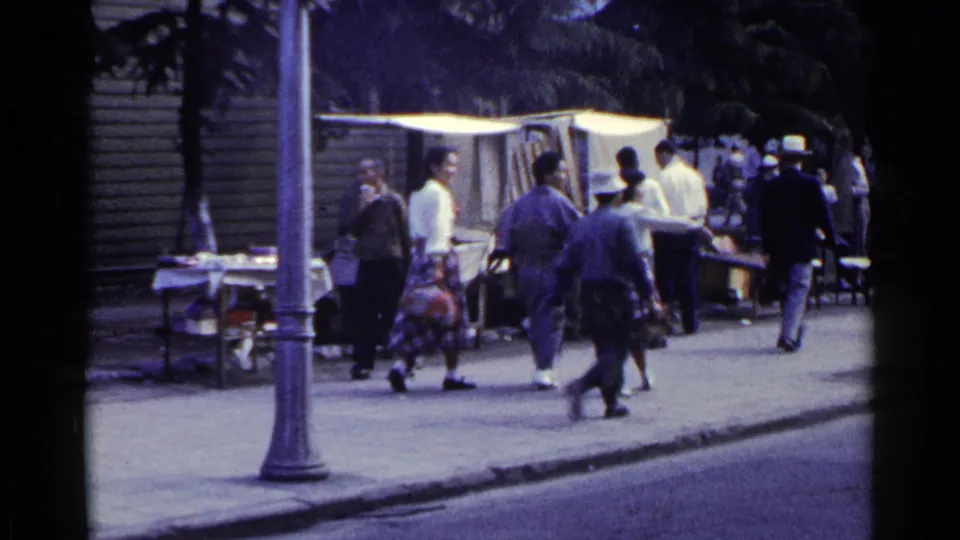 1953 Japan street frame with pedestrians walking by a lamppost, small vendor tables, and chairs set along the sidewalk.