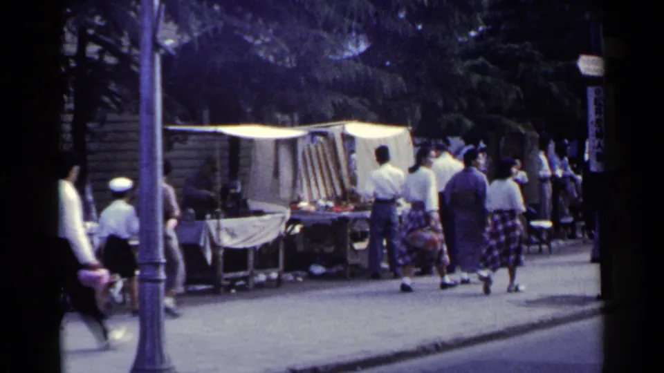 1953 Japan 8mm frame showing people in mixed traditional and Western-style clothing approaching a sidewalk stall area.