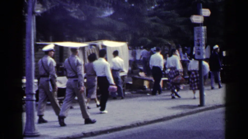 1953 Japan street frame with several pedestrians passing a canopy-covered sidewalk stall, chairs, and a vertical street sign.