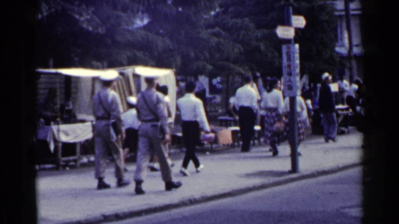 Two uniformed men walk along a sidewalk past market stalls and pedestrians.