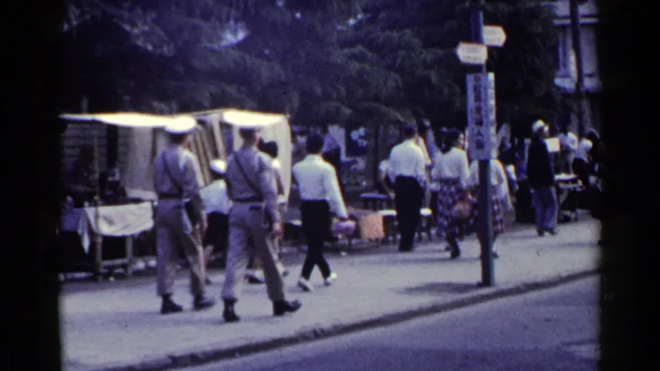 1953 Japan street view with two uniformed men walking past white-canopied stalls, pedestrians, signage, and a roadway.