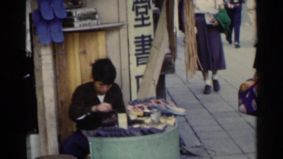 1953 Japan sidewalk frame showing a seated shoe repairman, displayed shoes or repair goods, vertical signage, and passing pedestrians.