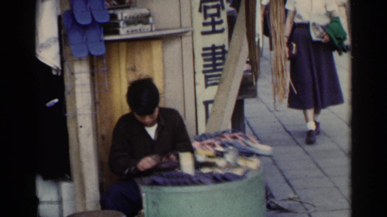 Seated shoeshiner works at a small street stall with shoes, slippers, and vertical signage nearby.
