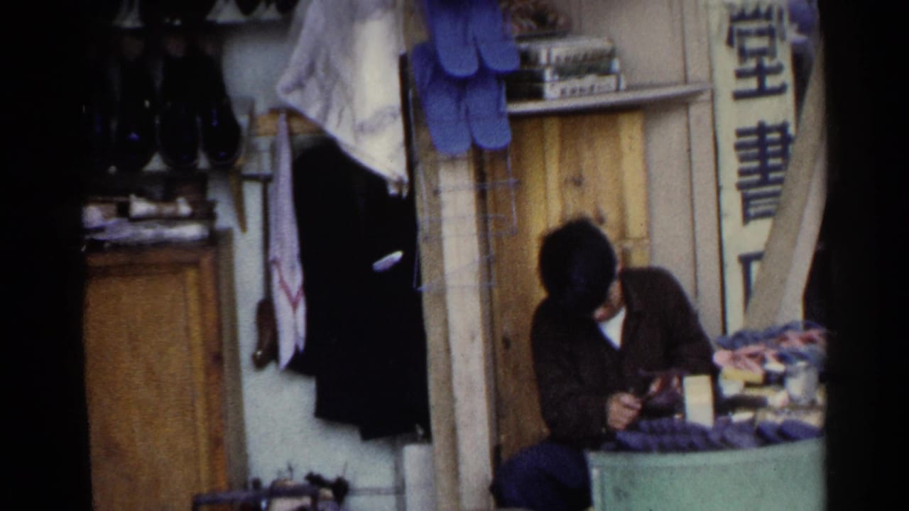 Shoeshiner continues working beside a wooden counter with footwear and hanging cloths behind him.