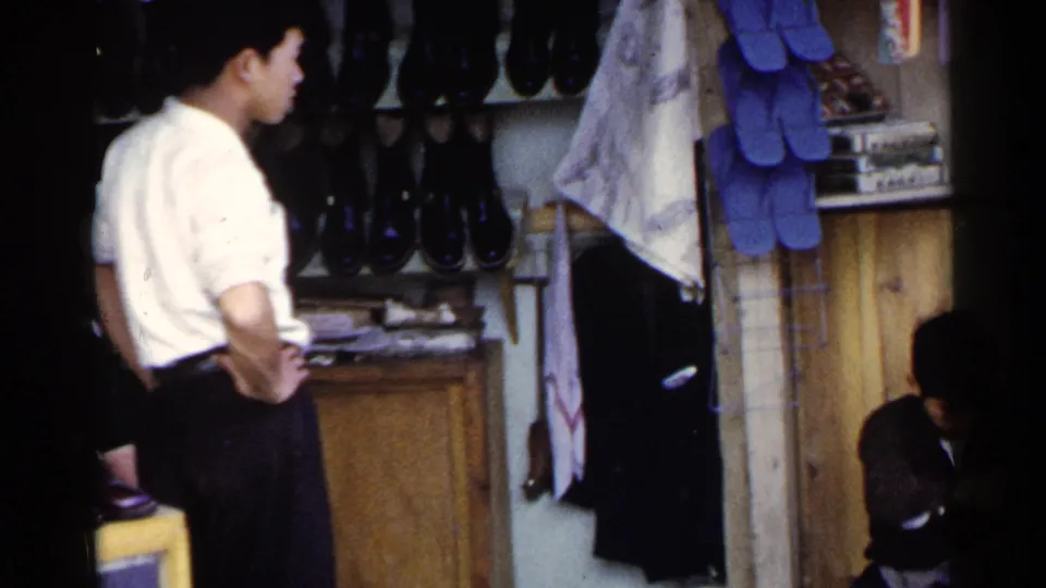 A waiting customer stands beside a Japanese street shoeshine stall as the seated worker remains partly visible near the lower frame.