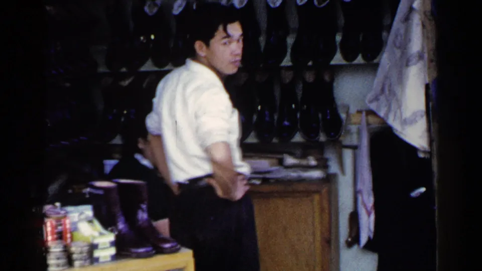 The customer turns toward the camera at the shoeshine stall, with footwear shelves, a wooden counter, and the seated worker nearby.