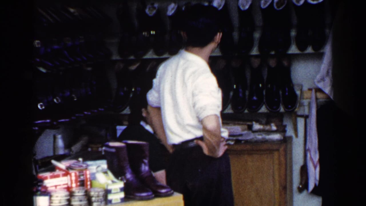 Man in a white shirt stands inside a dim shoe shop beside shelves of footwear.