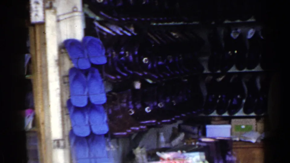 Blue sandals hang on the wall beside crowded shelves of dark footwear in the Japanese shop interior.