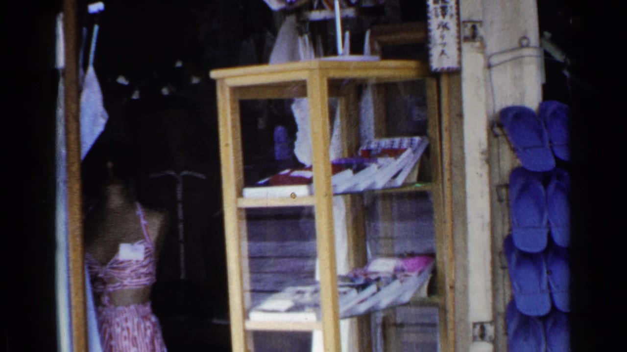 1953 Japan 8mm frame showing a storefront display case, hanging blue footwear or goods, and a dark shop entrance.