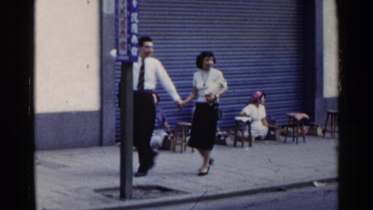 1953 Japan street scene with a couple walking past three women seated on small stools near a blue corrugated shutter.