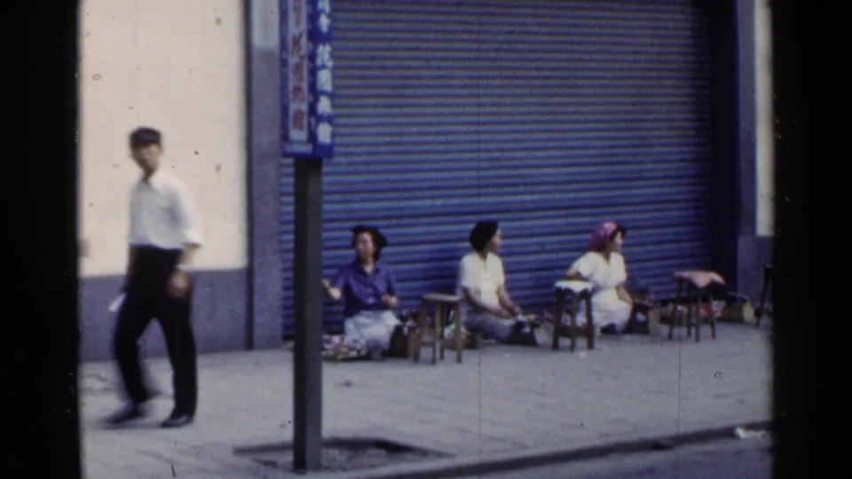 1953 Japan 8mm frame with a capped pedestrian crossing the foreground while women sit outside a shuttered storefront.