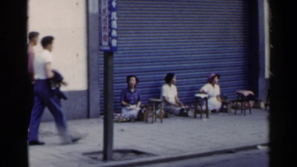 1953 Japan street frame showing passing men in the foreground and women seated along the sidewalk by a blue shutter.