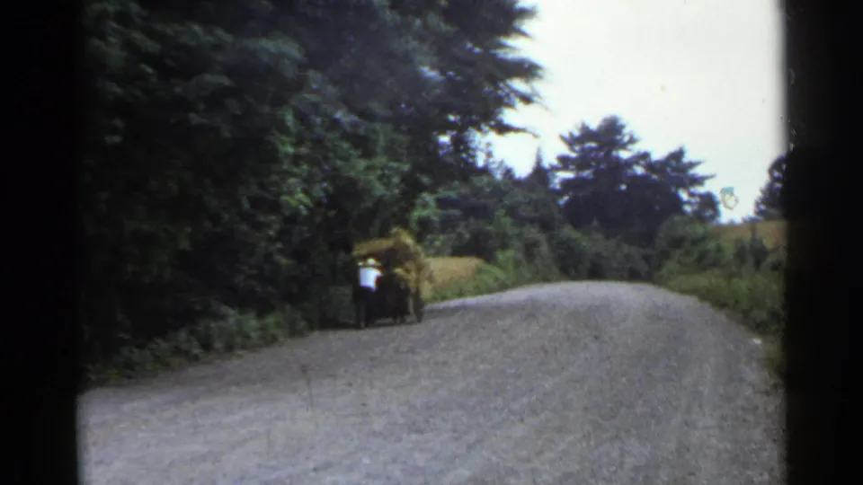 A distant man pushes a loaded cart along a winding rural road, captured with soft color and visible 8mm frame edges.
