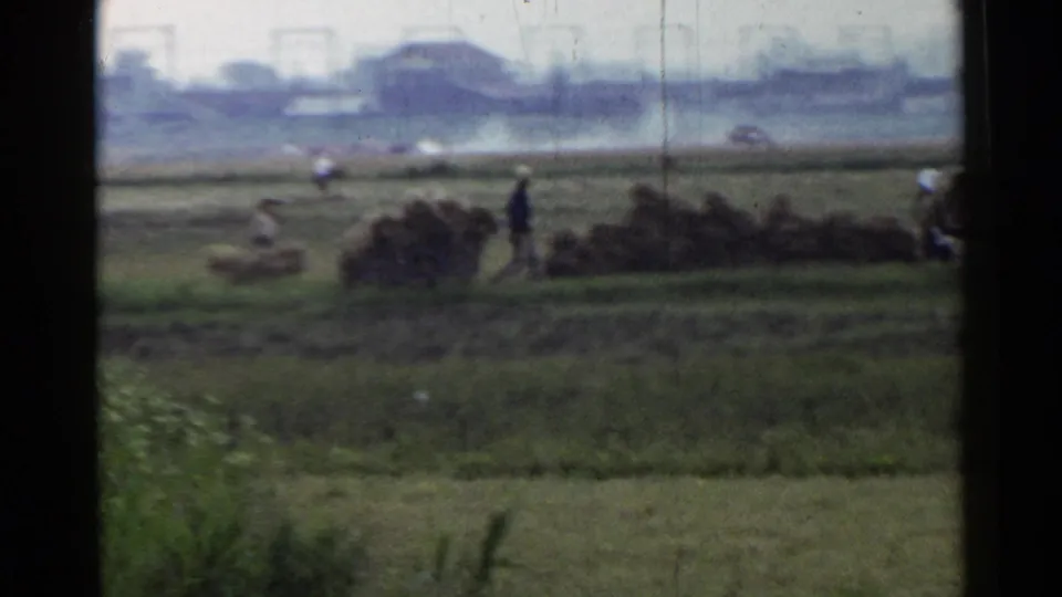 A soft 8mm view of rural Japan shows workers near grain piles, with hazy industrial structures in the distance.