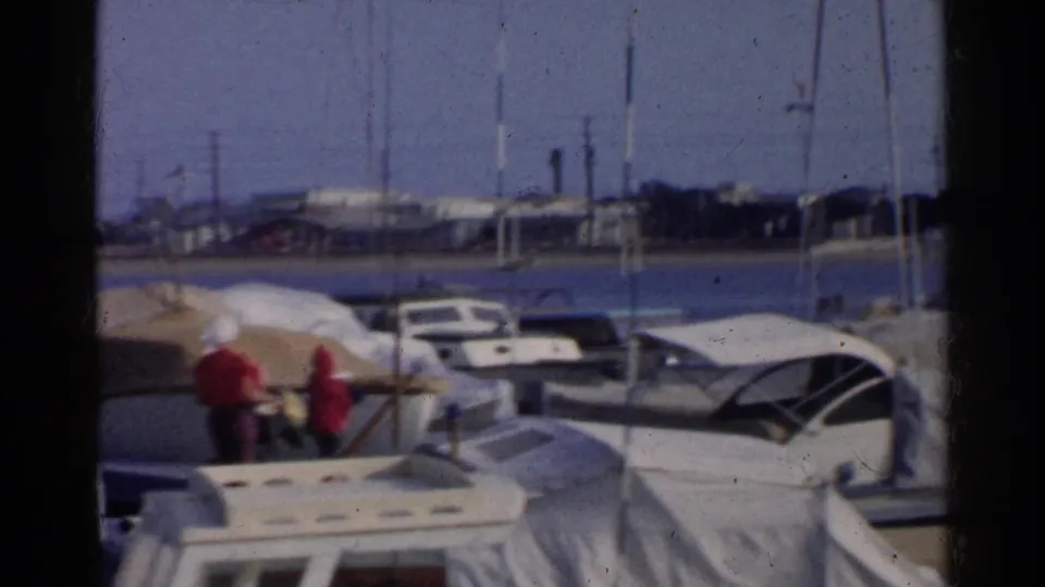 Two people in red shirts move along a California marina dock in 1955, surrounded by covered boats, masts, and harbor water.