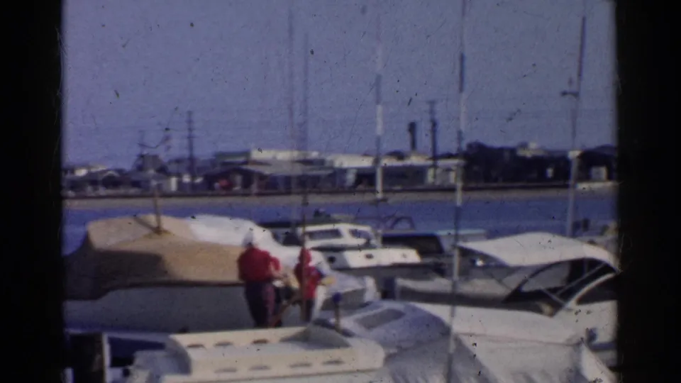 In a 1955 California harbor, two red-shirted people walk along a dock lined with covered motorboats and sailboat masts.