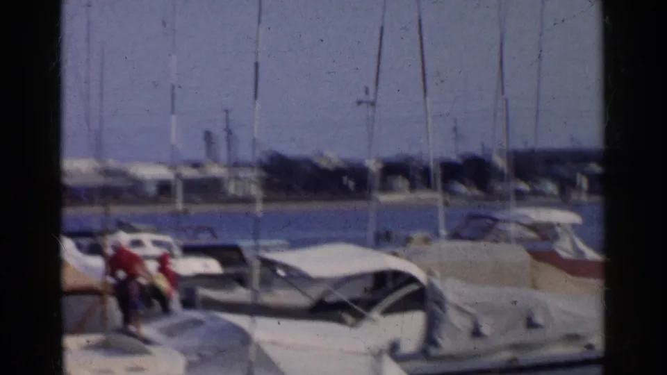 Soft 8mm frame of two people on a California marina dock in 1955, with covered boats, masts, water, and distant shoreline structures.