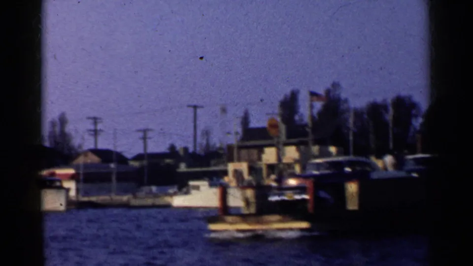 The camera frames a marina shoreline with a small boat in the foreground, low buildings, trees, and poles under a clear sky.