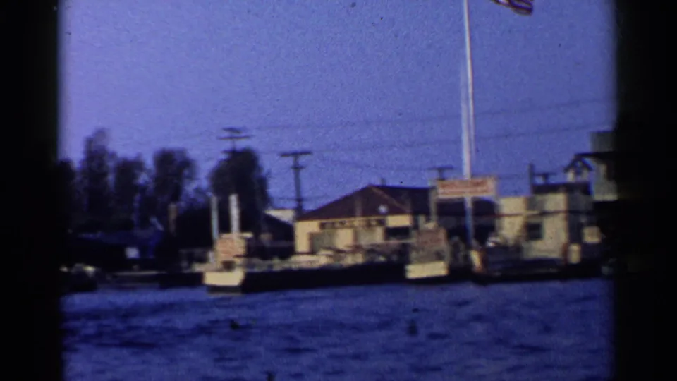 1955 California 8mm frame of a riverside or waterfront edge with buildings, flagpoles, utility lines, trees, and water in the foreground.