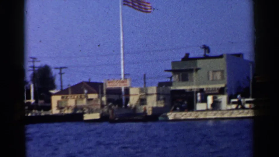 1955 California boat-view frame with a large American flag, low waterfront buildings, utility poles, background trees, and blue water.