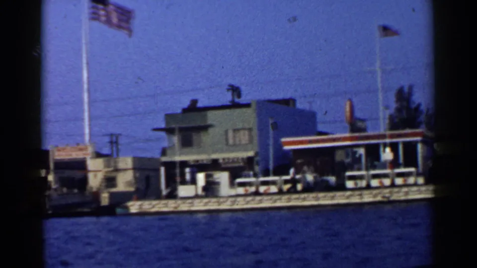 1955 California 8mm frame showing a waterfront building, tall American flagpole, railing or dock edge, small flag, and water from a boat.