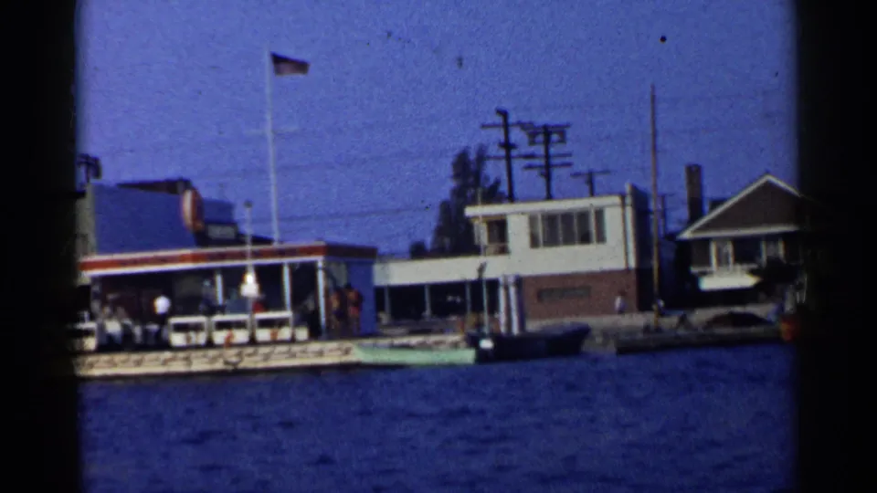 California waterfront frame showing a service-station-like harbor building, American flags, utility wires, docked boats, and a nearby house.
