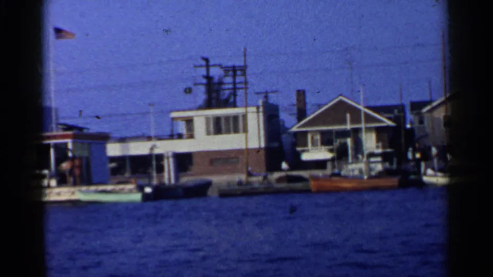 1955 California waterfront view with small boats at the dock, low buildings, American flags, overhead wires, and people near the harbor edge.