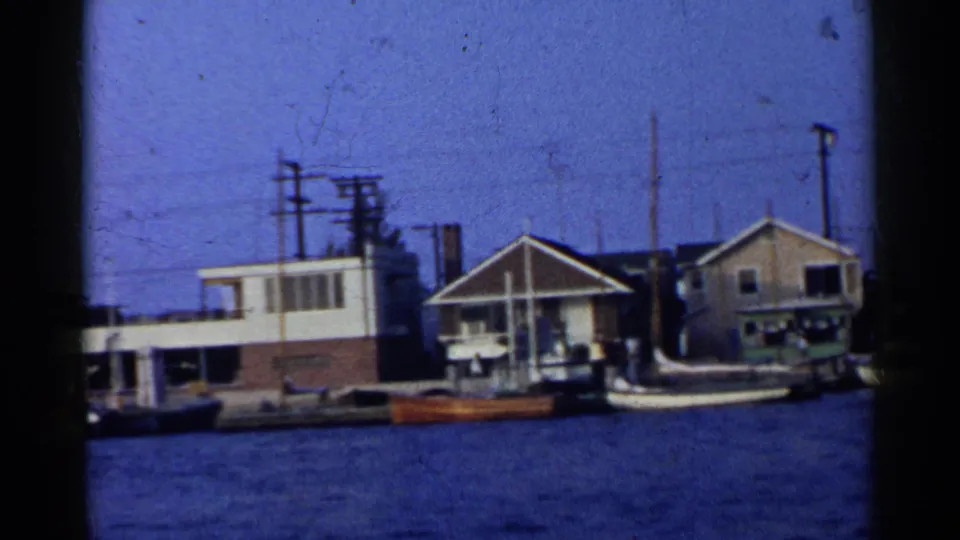 California harbor frame with waterfront houses, several docked boats, utility poles and wires, flags, and rippled blue water in front.