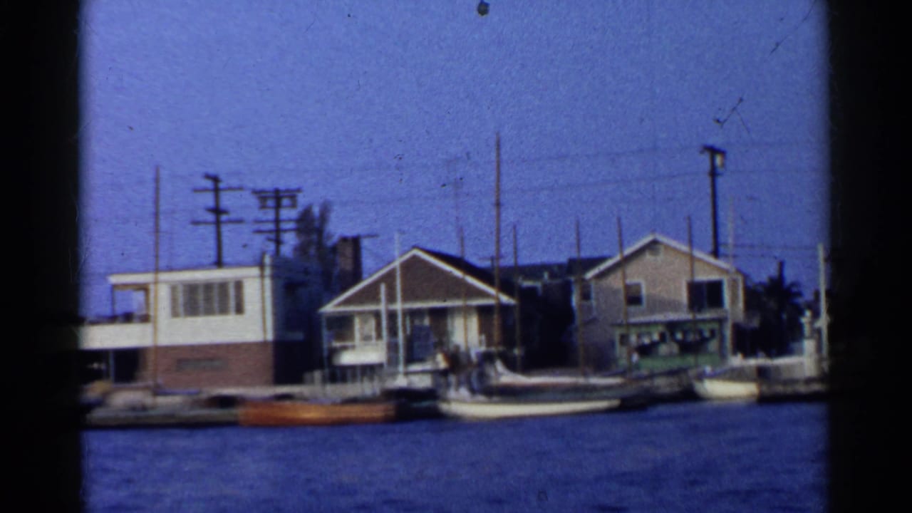 1955 California 8mm view of modest waterfront houses, docked boats, utility poles, and calm water.