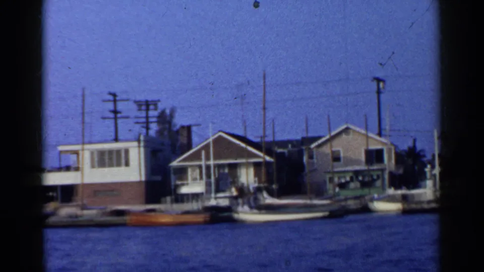 1955 California 8mm view of modest waterfront houses, docked boats, utility poles, and calm water.