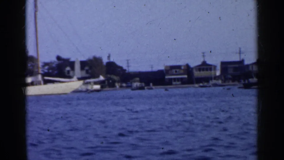 1955 California waterfront scene with a large white sailboat, shoreline homes, trees, and utility poles.