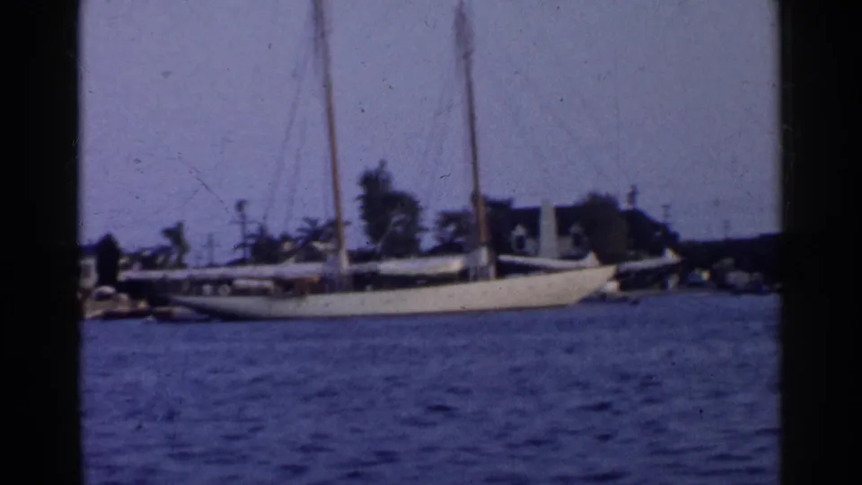 1955 California home-movie frame showing a white sailboat on calm water with waterfront buildings and trees behind it.