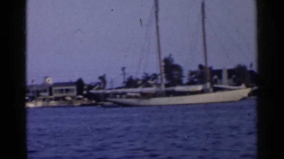1955 California 8mm frame showing rippled harbor water, a white sailboat, shoreline houses, and palms through a dark film-frame edge.